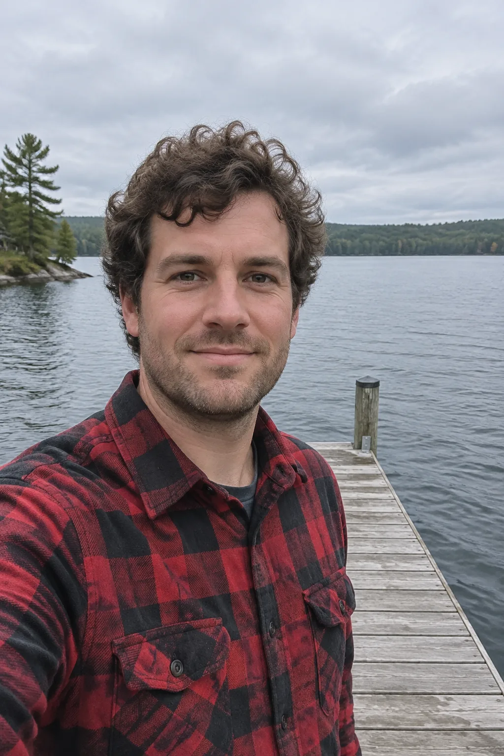 Man with curly brown hair and stubble in a red plaid flannel shirt, on a lakeside dock