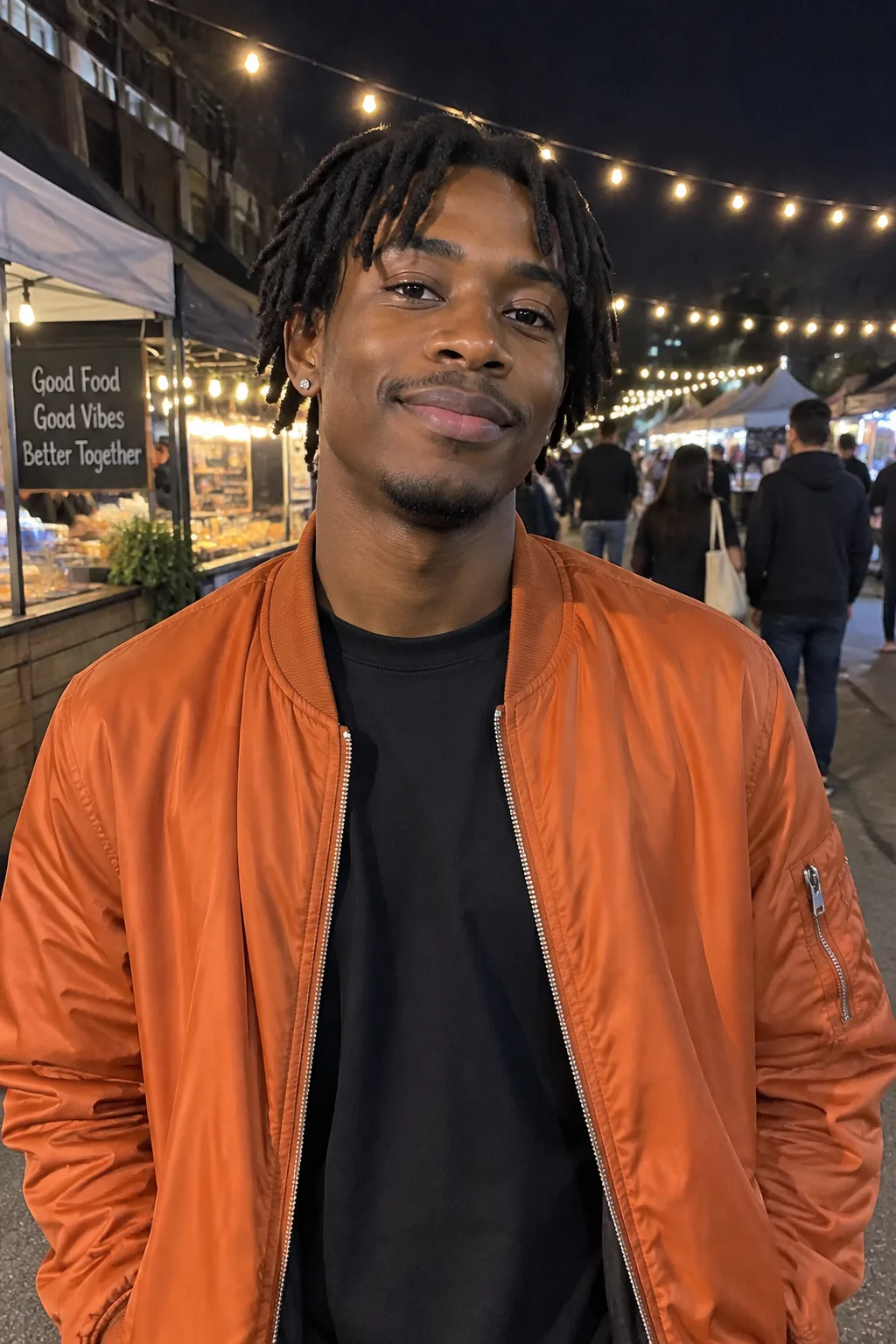 Joven con rastas cortas y una chaqueta bomber naranja, en un mercado nocturno con guirnaldas de luces