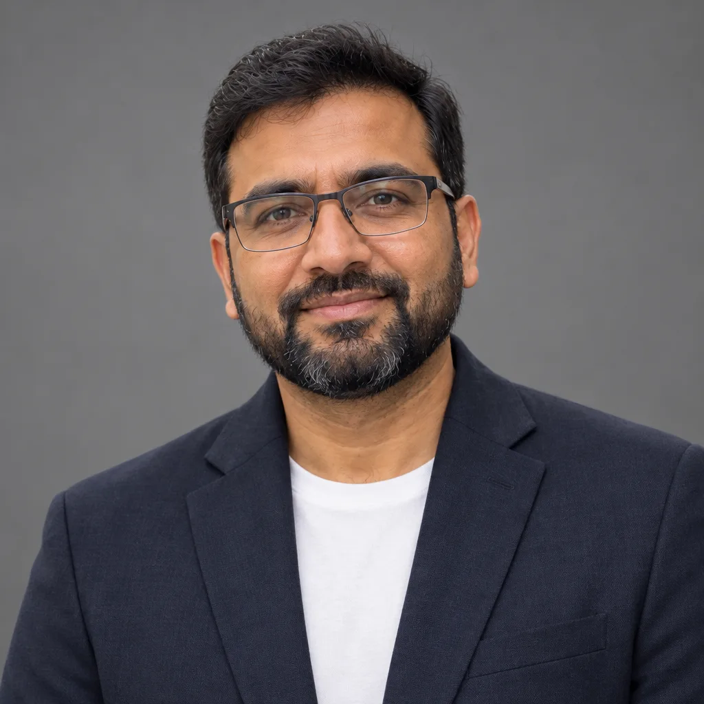 The same man in a corporate headshot: a dark blazer over a white tee against a plain grey studio backdrop