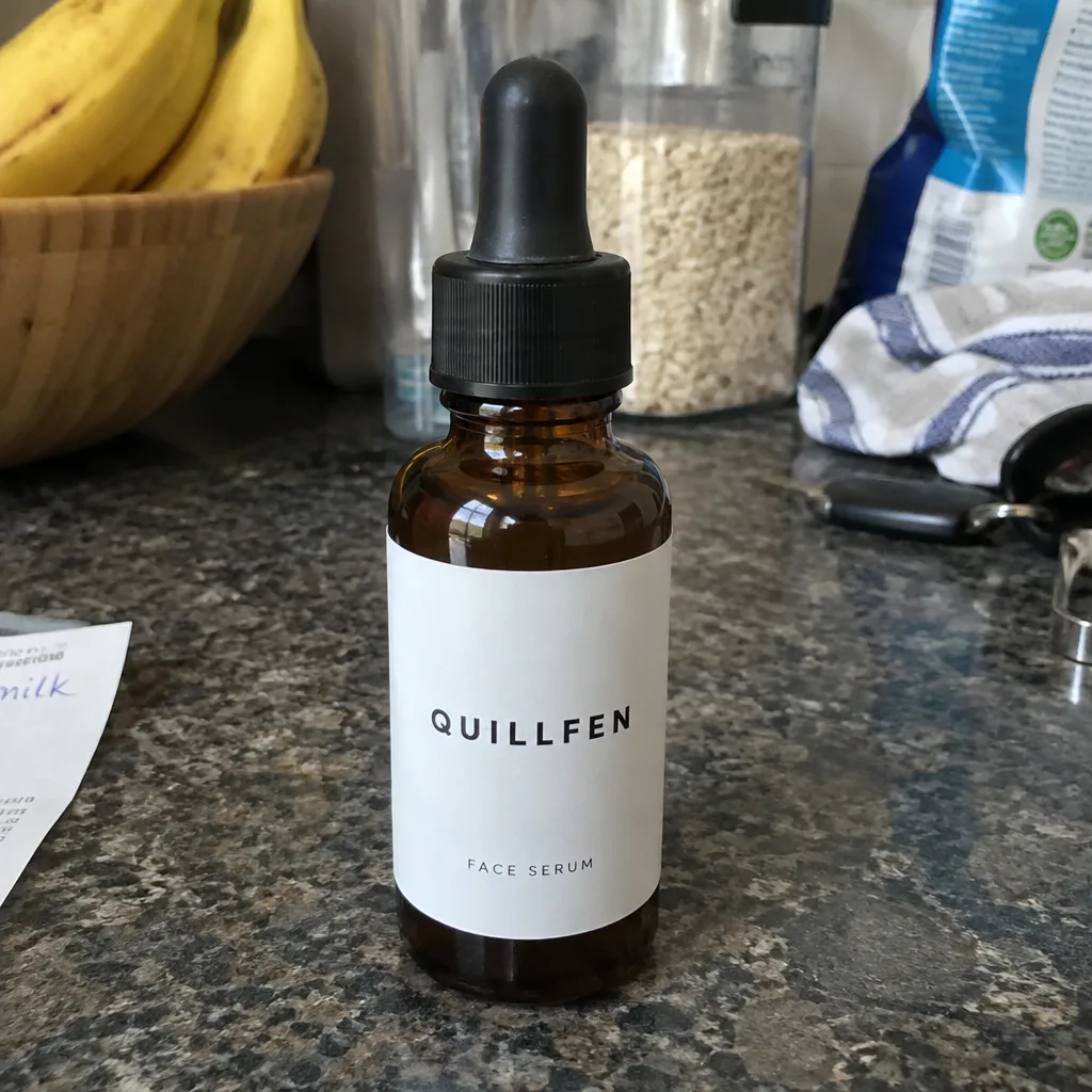 Amber glass dropper bottle with a white label reading QUILLFEN FACE SERUM on a cluttered kitchen counter