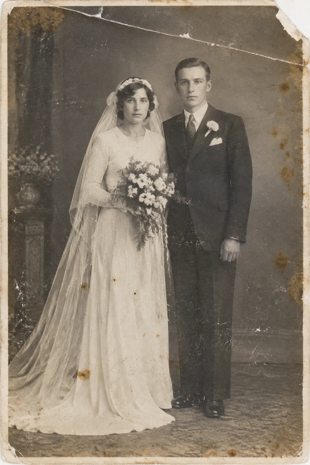 Faded, scratched 1930s black and white wedding portrait of a bride in a long white dress and veil holding a bouquet beside a groom in a dark suit, with a torn corner and brown stains