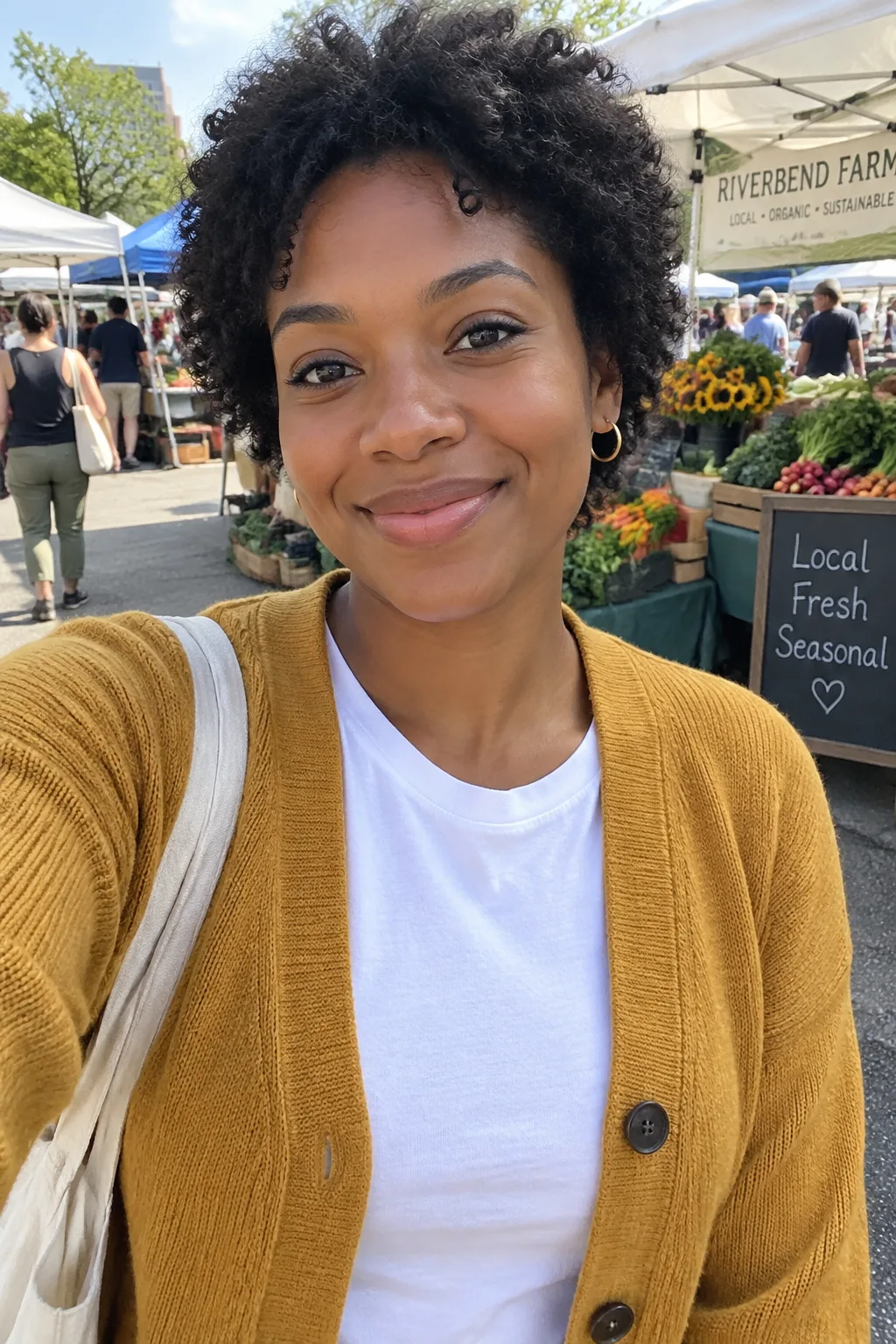 Young woman with short natural curls in a mustard cardigan, smiling at an outdoor farmers market