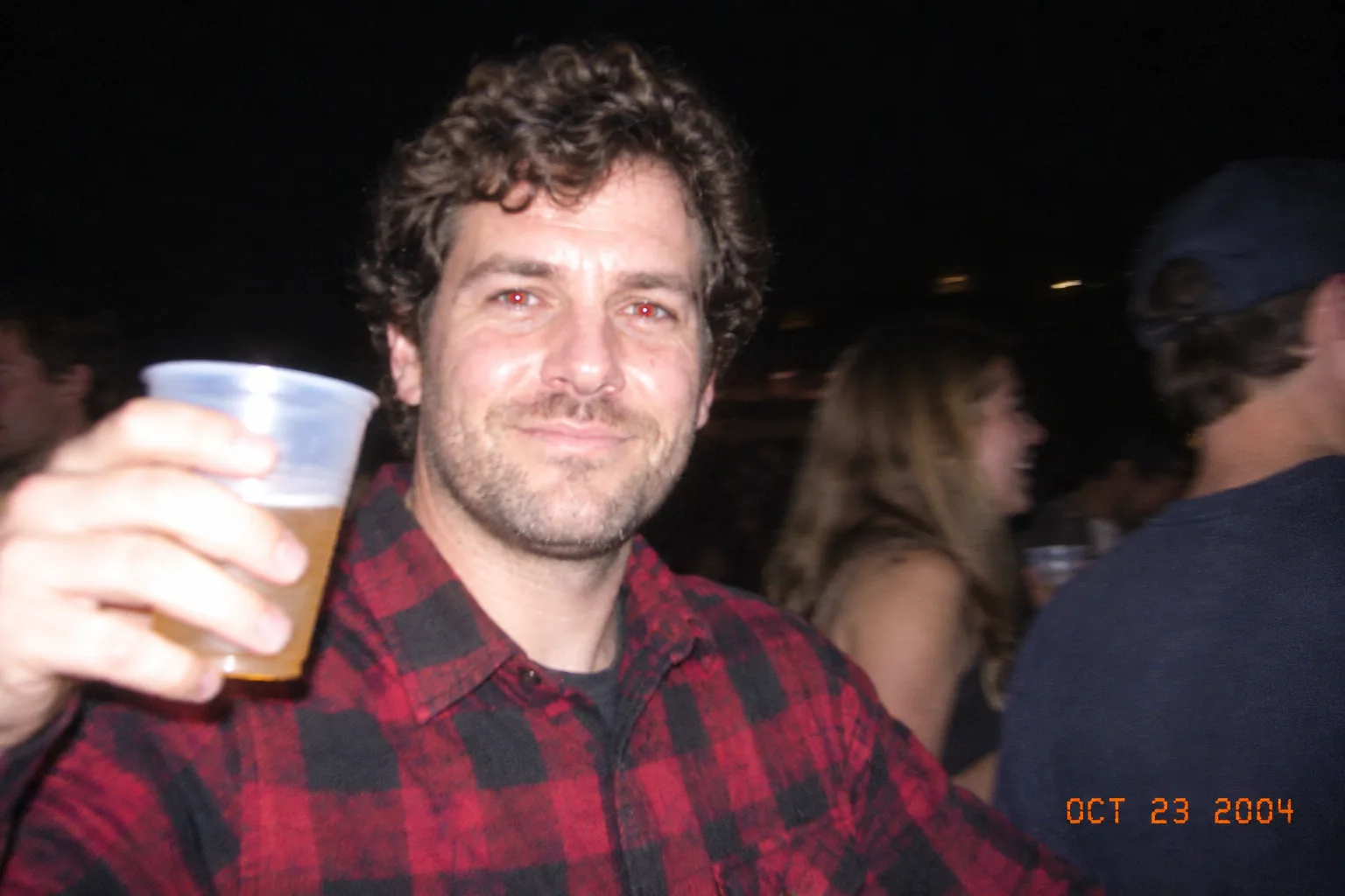 The same man raising a plastic cup at a night party, lit by direct flash, with an orange date stamp